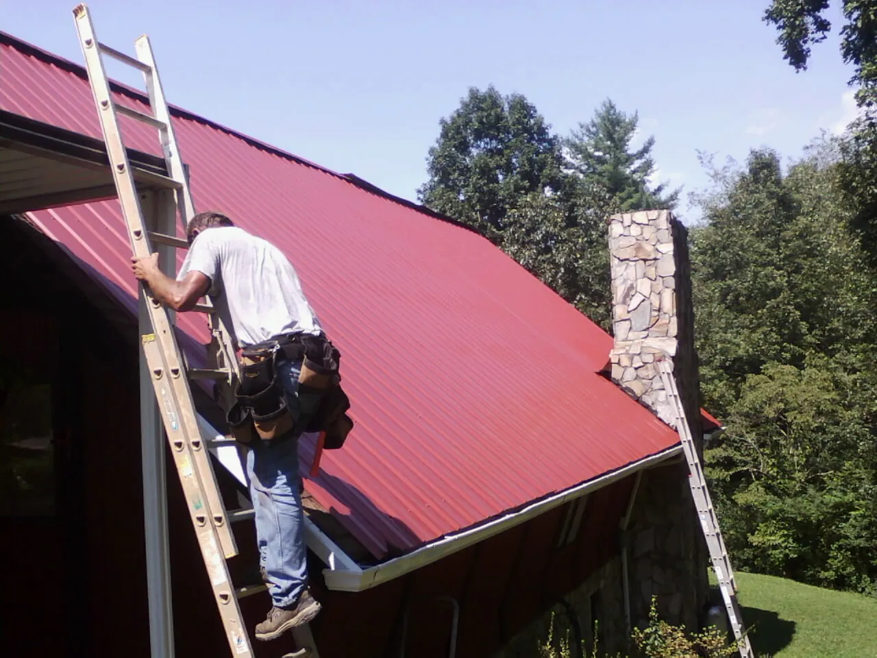 Roofer on a ladder beside a red metal roof and stone chimney — Soffit & Fascia Repair by Roofing Brothers in Italy